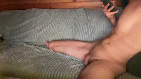 Video of a nude man, fair-skinned, with an erect penis, lying on a bed with a gray, ribbed blanket. He holds a green smartphone in his right hand, taking a selfie.