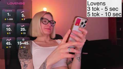 A video of a blonde woman with glasses, tattoos, and a white top, holding a phone. She's sitting in a dimly lit room.