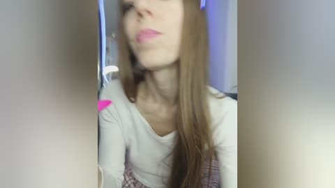 Video of a young woman with long brown hair, wearing a white long-sleeve shirt, making a pouty face, blurred background.