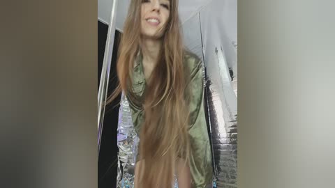 Video of a smiling young woman with long, straight, light brown hair, wearing a loose, olive-green blouse, standing indoors near a partially open door, with a brick wall and white ceiling visible in the background.