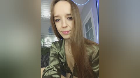 Video of a young woman with long brown hair, fair skin, and light makeup, wearing a green satin blouse, seated indoors. Background features a white door and a glass jar.