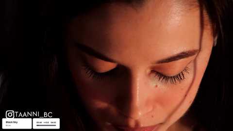 A close-up video of a woman with closed eyes, her skin glistening under warm light. Dark hair frames her face. The background is dark, emphasizing her features. Text in the bottom left reads \"@TAANNIBC\" and \"MUAHOF 2020\".