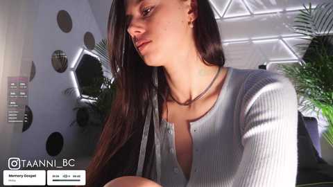 Video of a fair-skinned woman with long dark hair, wearing a light gray ribbed sweater, sitting indoors near hexagonal patterned wall.