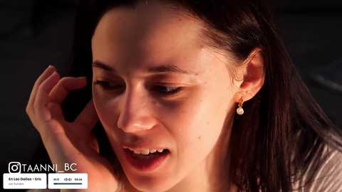 Video of a young woman with long dark hair, fair skin, and delicate features, wearing a silver necklace, talking on a phone, with a blurred background.