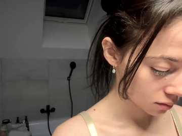 Video of a young woman with fair skin, dark hair in a bun, wearing a beige tank top, standing in a modern bathroom with white tiles, a showerhead, and a window.