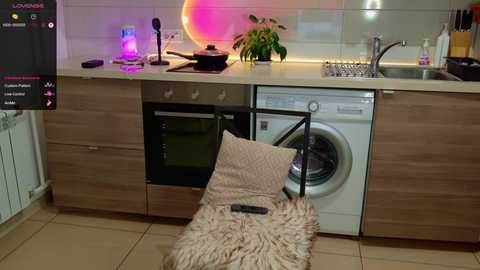 Video of a modern kitchen with wooden cabinets, a washing machine, a hanging drying rack, a potted plant, a remote control on a fur rug, and a colorful LED strip lighting the backsplash.