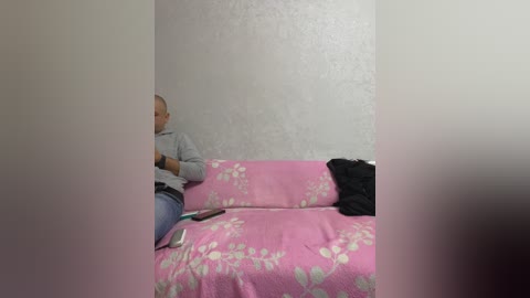 A video shows a bald, middle-aged man in a gray sweater and jeans, seated on a pink couch with white floral patterns, facing a gray, textured wall. A black jacket lies on the couch.