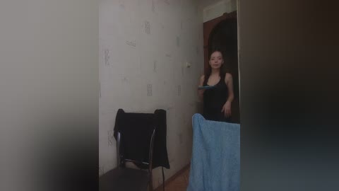 A video depicts a young woman with long hair, wearing a black tank top, standing in a dimly lit hallway. She holds a blue towel, with a black chair and a worn-out wall in the background.