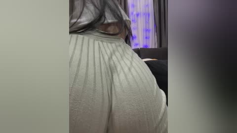 A video of a woman's lower back and buttocks in light grey, ribbed leggings, captured from behind. Background includes a dark couch and purple-lit wall.