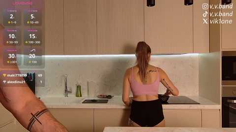 A video of a woman in a light pink sports bra and black shorts, cooking in a modern kitchen with light wood cabinets and a white marble backsplash.