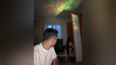 A video of a man with short dark hair, wearing a white T-shirt, seated indoors. A woman, partially visible, is in the background, wearing black lingerie, standing near a mirror with colorful light effects.