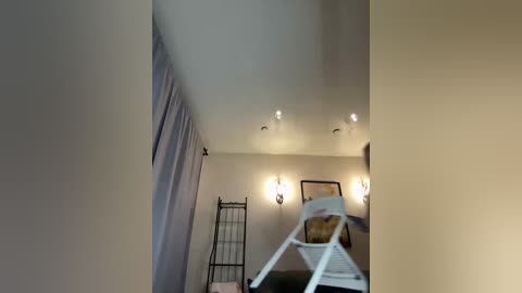 Video of a dimly lit, beige-painted bedroom with a white ladder chair, a white metal folding chair, and a light brown cushion. A white metal folding ladder stands beside the wall.