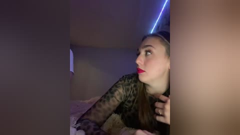 A video of a young woman with fair skin and red lipstick, wearing a camouflage-patterned long-sleeve shirt, lying on a bed, peering through a hole in a dark wall.