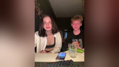 Video of a young woman with shoulder-length dark hair and a black bra, sitting next to a young man with curly hair, both smiling. They are indoors, in a dimly lit room with a gaming setup in the foreground.
