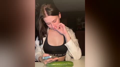 A video of a young woman with long dark hair, wearing a black sports bra and beige jacket, holding a cucumber, frowning, in a dimly lit room.
