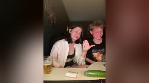 Video of two women, one with long dark hair, the other with curly blonde hair, laughing, sitting at a table with a beer, cucumber, and a T-shirt.