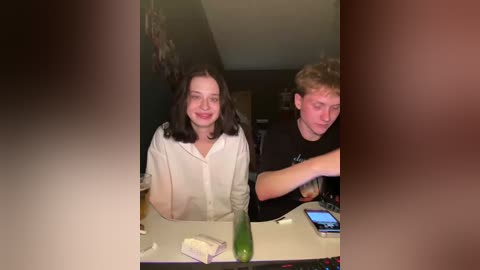 Video of two young people, a girl with shoulder-length dark hair and a boy with curly brown hair, sitting at a table. The background shows a dimly lit room with a green cucumber on the table.