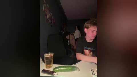Video of a young man with short hair wearing a graphic t-shirt, sitting at a table with a beer, cucumber, and chopsticks. Background shows dimly lit room with a wall art.