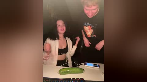 Video of two people in a dimly lit room. A woman with curly hair and a white robe holds a large erect penis. A man with short hair and a t-shirt stands beside her. A green cucumber sits on a desk.