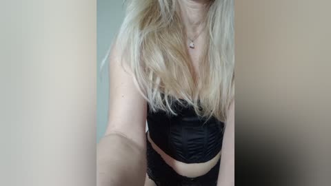 Video of a fair-skinned woman with long, blonde hair, wearing a black, lace-trimmed corset and black panties. The background is blurred, with soft, neutral tones.