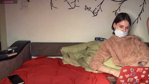 Video of a young woman with a surgical mask, wearing a brown sweater, lying on a bed covered in red and green blankets. The background shows a beige wall with black abstract branches.