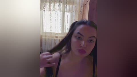 Video of a young woman with light skin, long dark hair, and pink lipstick, pulling a curtain aside in a dimly lit room with a wooden wall and lace curtains.