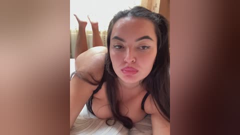 Video of a young woman with long dark hair, fair skin, and full lips, wearing a black bra, lying on a bed with striped sheets. The image is cropped with blurred foreground and background.