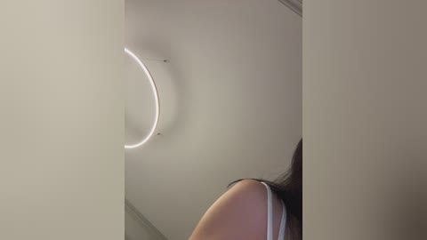 Video of a woman with long, dark hair, wearing a white top, standing in a modern, minimalist room with a white ceiling and circular LED light.