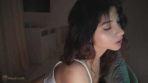 A video captures a young woman with long, wavy dark hair, wearing a white tank top, sitting indoors with a dimly lit background.