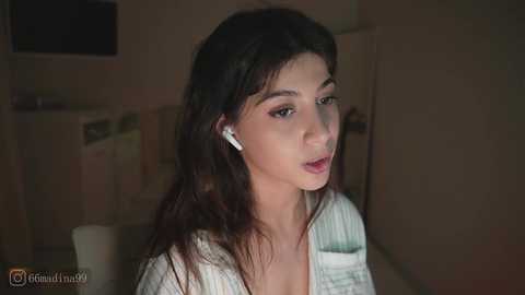 A video of a young woman with long dark hair, wearing a white shirt, listening to white earbuds in a dimly lit room with beige walls.