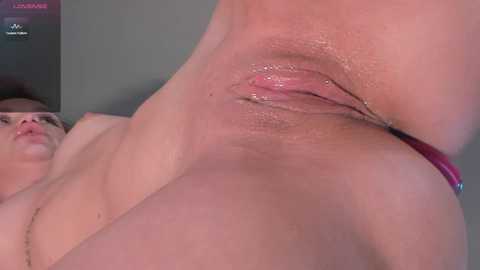 Close-up video of a nude woman with fair skin and visible labia, lying on her back, with a dark-haired man in the background.