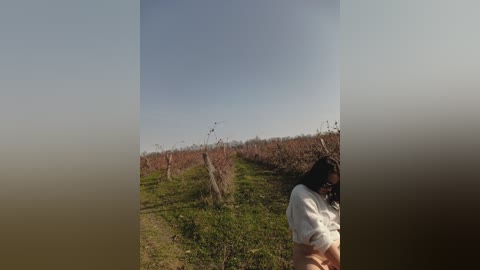 Video of a young woman with long dark hair, wearing a white sweater and pink shorts, standing in a vineyard under a clear blue sky.