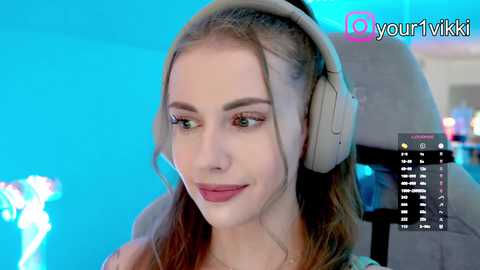 Video of a young woman with fair skin, light brown hair, wearing a headset, smiling softly. Background is a vibrant blue and grey, with a social media username \"yourviki\" and a chat interface on the right.