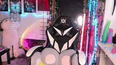 Video of a gamer in a black, white, and pink gaming chair with a plush dog, neon lights, and colorful toys in a cluttered, cozy room.