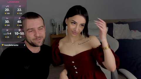 A video of a bearded man and a slender woman with black hair in a dark red dress, smiling in a modern bedroom with a bed and nightstand.