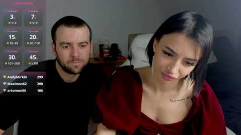 Video of a man and woman in a dimly-lit bedroom, both wearing casual clothes. The woman, with dark hair and tattoos, looks sad.