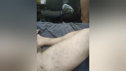 Video of a nude person lying on a gray bedspread, with visible pubic hair and a hand touching their genitals. Background shows a black office chair and dark wooden furniture in a dimly lit room.