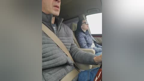 Video of a man in a grey quilted jacket and woman in a black puffer jacket, both wearing seatbelts, sitting in a car with beige seats.