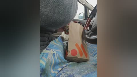 Video of a person wearing a grey hat, lying on a blue-patterned blanket in a car. A brown paper bag with red and orange markings is placed on the person's chest. The car interior is visible in the background.