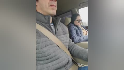 A video shows a man and woman in a car, both wearing winter jackets. The woman, with dark hair and glasses, looks pensive. The man, with a light beard, gazes straight ahead.