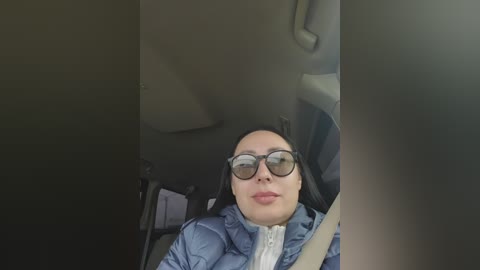 Video of a woman with long dark hair, wearing black-rimmed glasses and a blue jacket, sitting in a car, seen from the driver's seat.