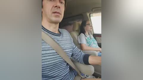 Video of a man wearing a striped shirt and a seatbelt in a car, next to a woman with glasses and a teal scarf, looking out the window.