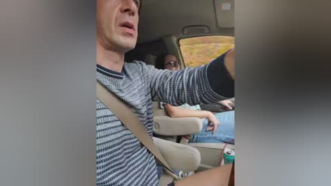 Video of a man in a striped shirt driving a car, with a woman in the passenger seat; both appear relaxed. The background shows a blurred view of a greenish-brown field.