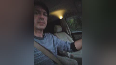 Video of a middle-aged man with dark hair, wearing a striped blue shirt, driving a car with a beige seat. The image is slightly blurred and has a muted color palette.
