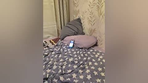 Video of a messy bedroom with a blue phone on a gray star-patterned blanket. A gray pillow is partially visible against a beige, leaf-patterned wallpaper.