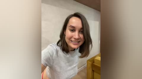 Video of a smiling young woman with shoulder-length dark brown hair, wearing a light gray T-shirt and pink shorts, standing in a small, brightly lit room with beige walls and a wooden dresser.