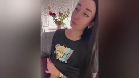 Video of a young woman with long black hair, fair skin, and a slim physique, wearing a black graphic T-shirt. She sits on a gray couch in a cozy room with floral wallpaper and a vase of flowers.