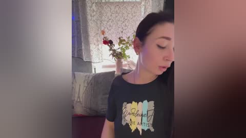 Video of a young woman with fair skin, dark hair, and light makeup, wearing a black T-shirt with a colorful design. She stands in a cozy room with lace curtains and a vase of flowers.