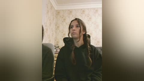 Video of a young girl with long brown hair in pigtails, wearing a black hoodie, seated on a gray couch in a dimly lit room with floral wallpaper and a framed picture on the wall.