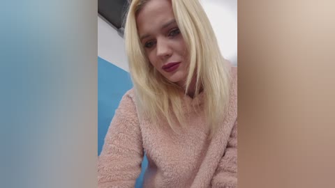 Video of a fair-skinned, blonde-haired woman with red lipstick, wearing a pink, textured sweater. She stands in a blue and white bathroom, with a blurred background.